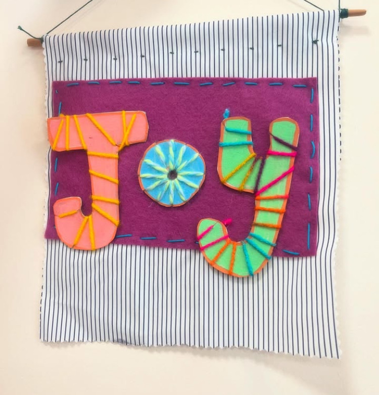 a fabric banner with a purple felt square sewn on in large stitches using brightly coloured yarn. Over this, the word 'joy' has been cut out of card, with each letter in a different bright colour, wrapped in yarn and sewn on.